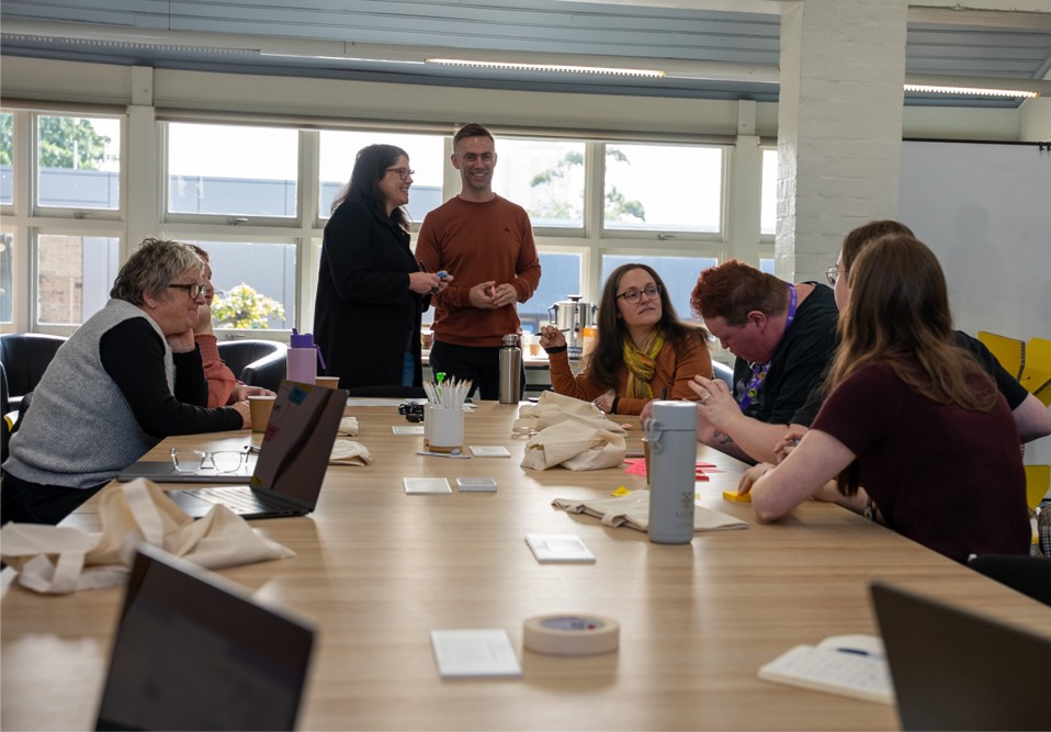 People sit and stand around a table in a meeting room, doing co-design.